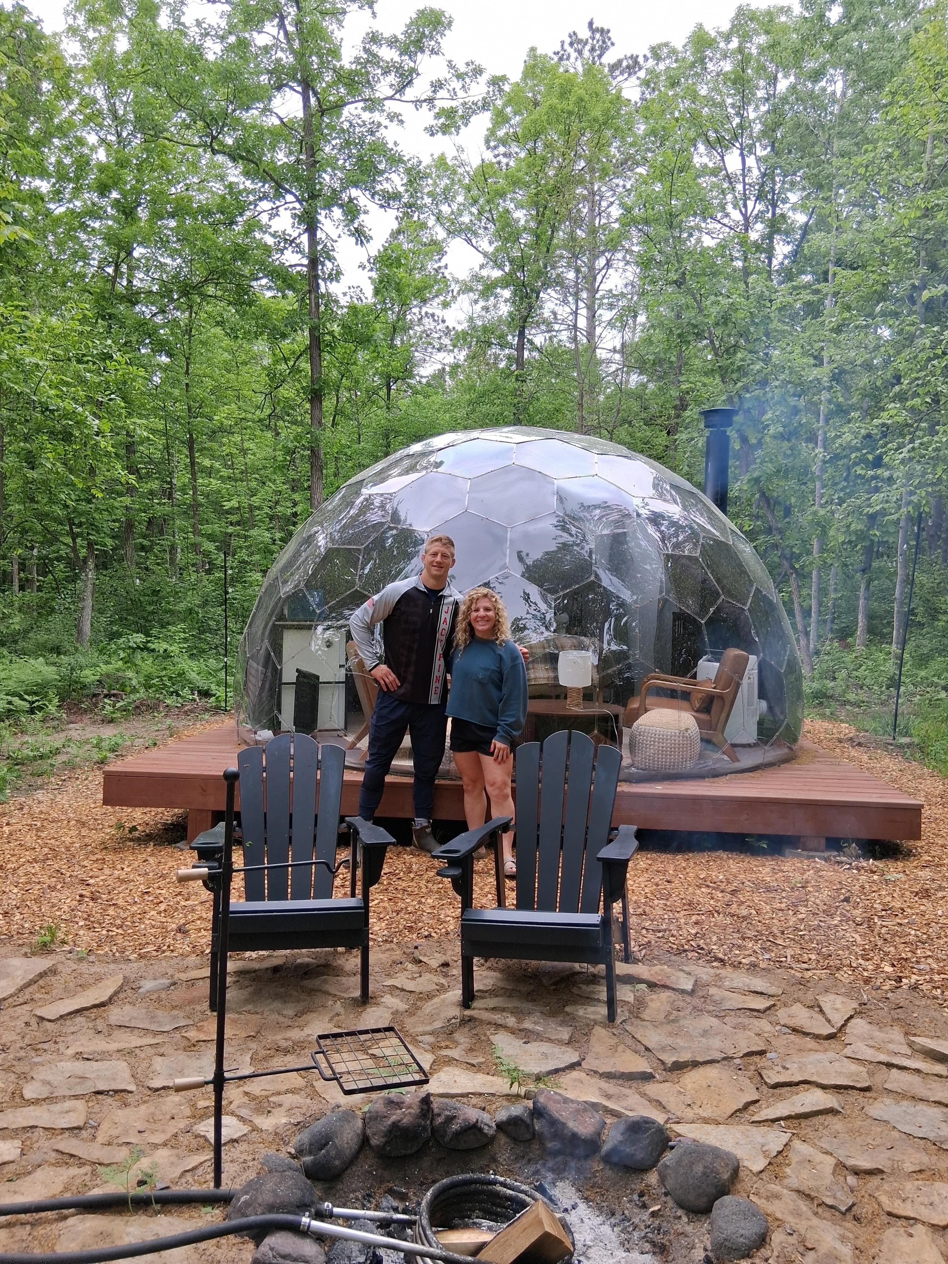 Two guests standing inside a geodesic dome with glass panels, fire pit and Adirondack chairs in front