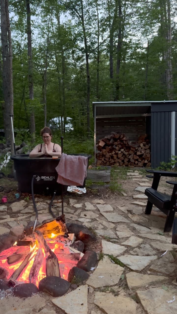 Person relaxing in a wood-fired hot tub at dusk surrounded by pine forest