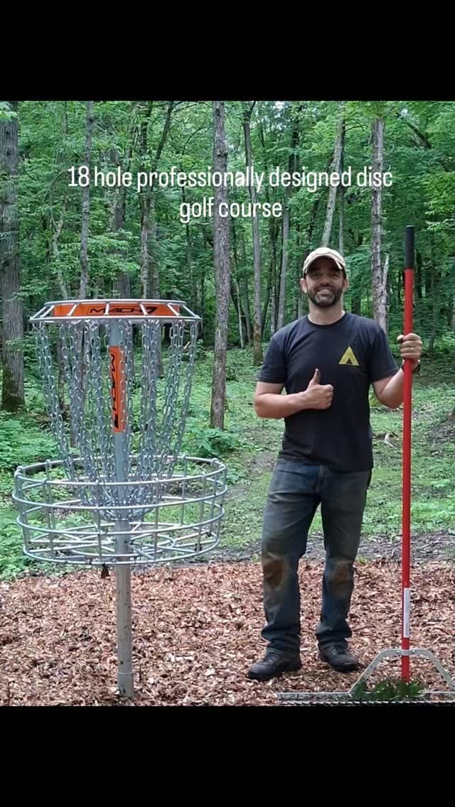 Person demonstrating disc golf with basket and wooded fairway behind